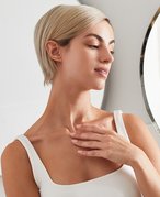 person with a blonde pixie cut wearing a white tank top and holding their hand to their neck