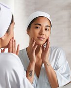 Person wearing a headband with their dark hair pulled back looking in the bathroom mirror and touching their cheeks