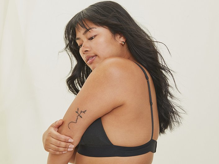 Person with long dark hair and bangs wearing a black bra and looking down at their tattooed arm while holding it