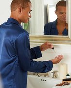Person in blue pajamas standing at the bathroom sink applying a skincare product to their hand with a dropper