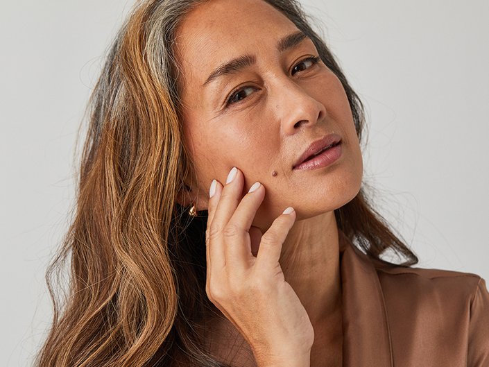 Person with long dark hair and brown eyes touching their fingers to their cheek