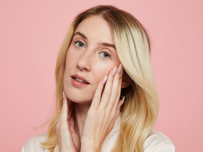 Picture of a blonde person touching the sides of their face against a pale pink background