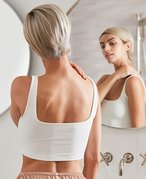 Person with a blonde pixie cut wearing a white tank top and looking in the bathroom mirror while touching their shoulder