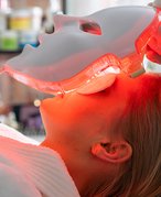 A person lying on their back in a white spa robe having an LED light mask placed on their face