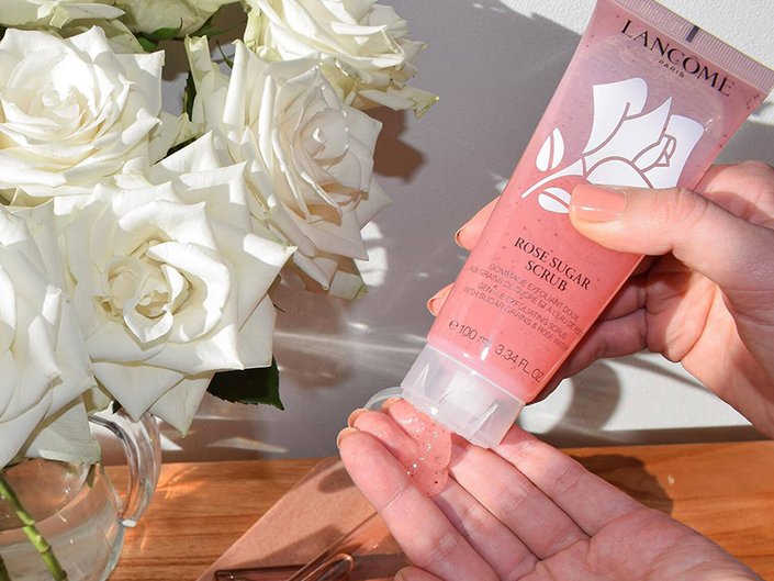 Person squeezing Lancôme Rose Sugar Exfoliating Scrub onto their fingers next to a vase of white roses