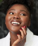 A person smiling and looking away from the camera touches their hand with red painted nails to their cheek