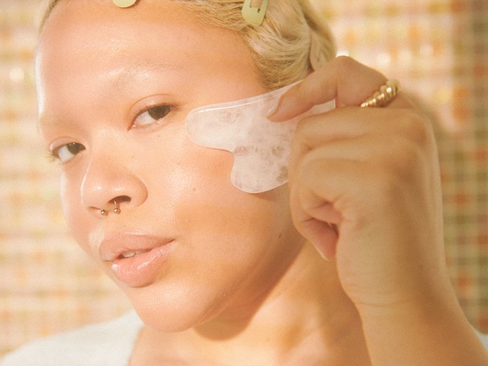 Close-up picture of a model with bleached eyebrows and a septum piercing, holding a white gua sha to their cheek in the sunlight 