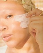 Close-up picture of a model with bleached eyebrows and a septum piercing, holding a white gua sha to their cheek in the sunlight 