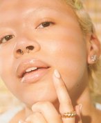 Close-up picture of a model with blonde hair and a septum piercing, touching the side of their mouth