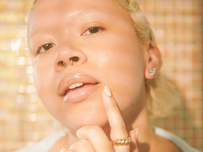 Close-up picture of a model with blonde hair and a septum piercing, touching the side of their mouth