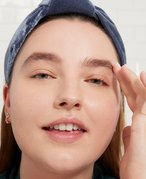 Close-up picture of a brunette model softly smiling and touching the side of their face, wearing a blue headband