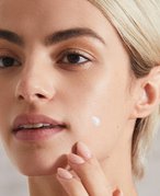 Person with a blonde pixie cut and a spot treatment on their cheek touching their chin with their finger tips